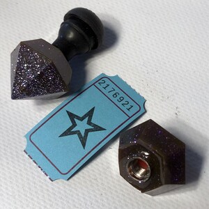 May include: Two brown and purple glitter resin bottle stoppers with a black knob on one. The stoppers have a hexagonal and diamond shape. A blue ticket with a star and the number 2176921 is in the background.