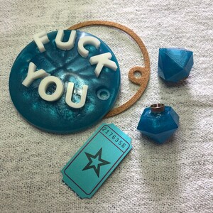 Può includere: Un sottobicchiere in resina blu con le parole "FUCK YOU" in lettere bianche. Inoltre, due oggetti geometrici in resina blu, un cerchio di sughero e un biglietto turchese con una stella e il numero 2176356.