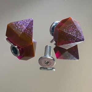 Può includere: Due tiretti per cassetti a forma di diamante in resina marrone e viola con glitter argentato incorporato nella resina. I tiretti sono fissati a viti argentate con una rondella argentata.