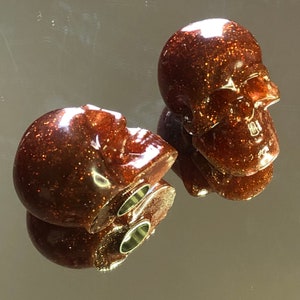 Peut inclure: Deux petites décorations en forme de crâne, de couleur marron et pailletées. Les crânes ont une finition brillante, presque métallique.