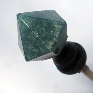 May include: A teal green, faceted, glitter-filled resin bottle stopper with a black base.