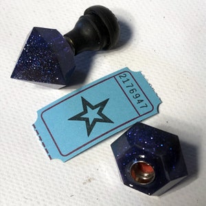 May include: Two blue glitter resin stamp tops with a black wooden handle. The stamp tops are shaped like a diamond and a hexagon. The handle is attached to the diamond-shaped stamp top. A blue ticket with a star and the number 2176947 is in the background.