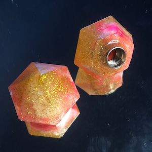 May include: Two pink and gold glitter resin bottle stoppers with a hexagonal shape. One stopper has a metal insert for a bottle.