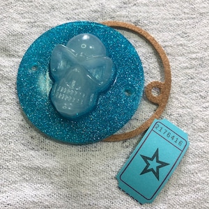 Peut inclure: Un crâne bleu pailleté avec un motif de crâne blanc. Le crâne est attaché à une sangle en cuir marron avec un ticket bleu attaché. Le ticket a une étoile et le numéro 2176416 imprimé dessus.