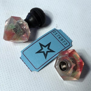 May include: A clear, faceted resin bottle stopper with red, green, and yellow glitter. A black rubber stopper is attached. A blue ticket with a star graphic and the number 2176973 is also present.