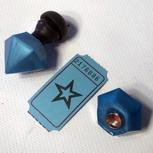 May include: Two blue glass bottle stoppers with a black knob on top. One stopper is shaped like a diamond and the other is a rounded hexagon. A blue ticket with a star and the number 2176886 is in the background.