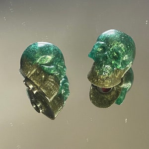 May include: Two green and gold skull-shaped bottle stoppers with a glittery finish.