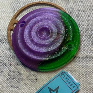 May include: A round, purple and green resin disc with a swirling, glittery pattern. The disc has a hole in the top and bottom.