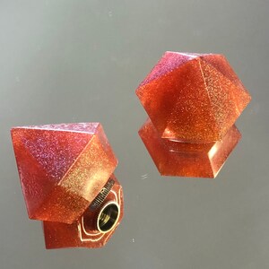 May include: Two sparkly, red, geometric bottle stoppers. One is a diamond shape with a metal insert and the other is a hexagonal shape.