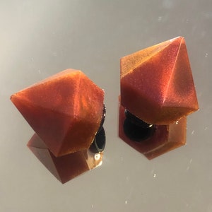 May include: A pair of iridescent red and gold resin earrings shaped like diamonds. The earrings have a shiny, glittery finish.