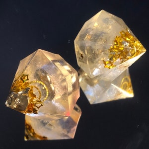 Peut inclure: Deux dés en résine géométriques transparents à facettes avec des paillettes dorées et des accents métalliques. Les dés ont la forme d'une pyramide avec un sommet pointu.
