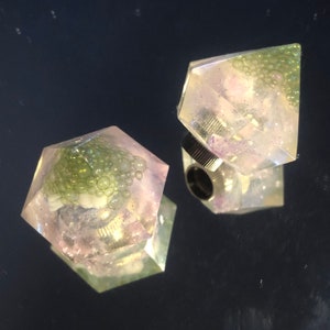 Peut inclure: Deux bagues géométriques en résine transparente et irisée. Chaque bague présente un amas de bulles vertes et un anneau argenté. Les bagues sont présentées sur une surface réfléchissante, créant un effet miroir.