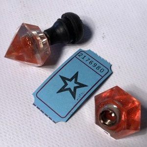Peut inclure: Deux bouchons de bouteille en verre facetté rouge et scintillant. Un bouchon a un dessus en caoutchouc noir. L'autre bouchon a un dessus en métal argenté. Un billet bleu avec le numéro 2176980 et une étoile noire est en arrière-plan.