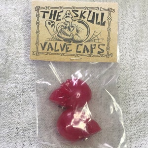 Peut inclure: Deux bouchons de valve en forme de crâne rouge emballés dans un sac en plastique transparent avec une étiquette en papier brun qui indique "The Skull Valve Caps".