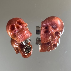 May include: Two reddish-brown skull-shaped plugs with a metallic silver centre. The plugs are split in half, with a magnetic connection. The skulls have a glittery, iridescent finish, and are displayed on a reflective surface.