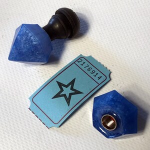May include: A blue, faceted resin bottle stopper with a dark brown handle, next to a blue ticket with a star graphic and the number 2176914. Another blue resin piece with a gold-colored metal insert is also present.