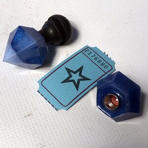 Peut inclure: Deux bouchons de bouteille en verre bleu, l'un en forme d'étoile et l'autre en forme de diamant, avec un bouchon de bouteille en bois noir en arrière-plan. Le bouchon en forme d'étoile a un ticket avec le numéro 2176880 dessus.