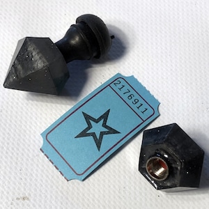 May include: Black, geometric bottle stoppers and a blue ticket with a star graphic. One stopper has a faceted, diamond-like shape. The ticket has the number 2176911 printed on it. The other stopper has a threaded interior.