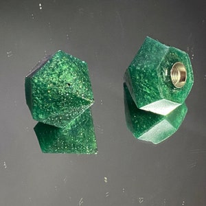 May include: Two green, geometrically shaped bottle stoppers with a speckled finish. One stopper has a threaded hole on the top.