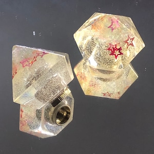 Puede incluir: Dos tapones de botella de vidrio transparente con un diseño de estrella. Los tapones tienen forma de diamante y pentágono. Los tapones tienen un diseño de estrella roja y blanca.