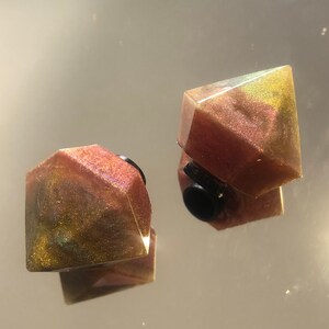 Puede incluir: Dos pendientes de resina geométricos e iridiscentes en tonos marrones, dorados y verdes. Los pendientes tienen un respaldo negro.