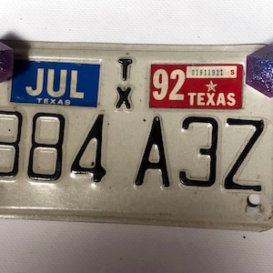 May include: A silver license plate with black lettering and numbers. The plate reads "JUL TEXAS 884 A3Z" and has a red sticker with a white star and the text "92 TEXAS".