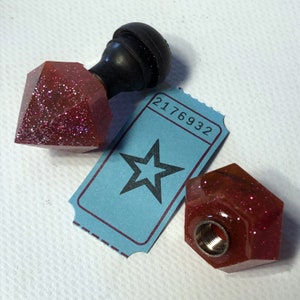 Puede incluir: Dos tapones de botella de resina roja con purpurina, uno con forma de diamante y el otro con forma de hexágono, con un mango de madera negro en la parte superior del tapón con forma de diamante. Un billete azul con el número 2176932 y una estrella negra en el fondo.