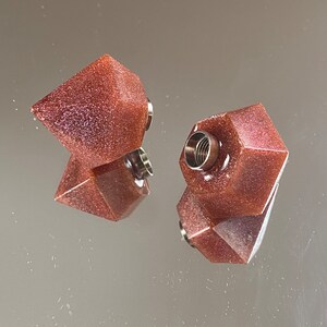 Può includere: Due tappi per bottiglie in resina rosso-marrone di forma geometrica con un top in metallo argentato.