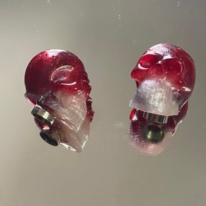 Puede incluir: Dos tapones de botella en forma de calavera de color rojo y blanco con anillos de metal plateado.