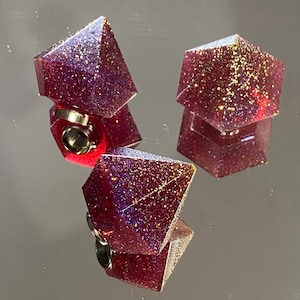 Könnte beinhalten: Drei rote, glitzernde Würfel aus Harz mit goldenen Flocken. Die Würfel sind in Form einer Pyramide, eines Würfels und eines Diamanten.