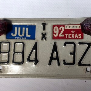 Peut inclure: Une plaque d'immatriculation argentée du Texas avec le numéro 884 A3Z. La plaque comporte un autocollant bleu et blanc qui dit "JUL TEXAS" et un autocollant rouge qui dit "92 TEXAS". Il y a deux formes géométriques rouges et scintillantes de chaque côté de la plaque.