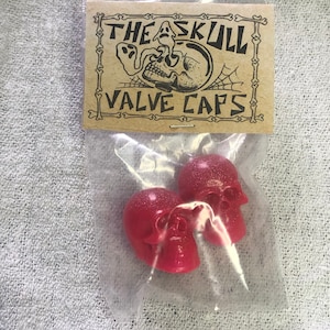 Può includere: Due tappi per valvole a forma di teschio rosso in un sacchetto di plastica trasparente con un'etichetta di carta marrone che recita "THE SKULL VALVE CAPS".