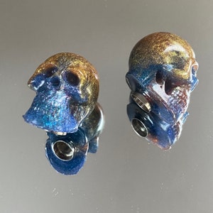 May include: Two blue and gold skull-shaped ear plugs with a silver metal base.