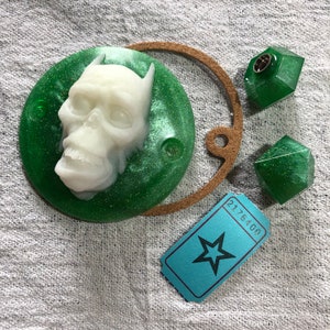 Peut inclure: Ensemble d'objets faits à la main. Un crâne blanc est posé sur un disque vert scintillant. Deux formes géométriques vertes, un anneau marron et un billet bleu avec une étoile et le numéro 2176400.