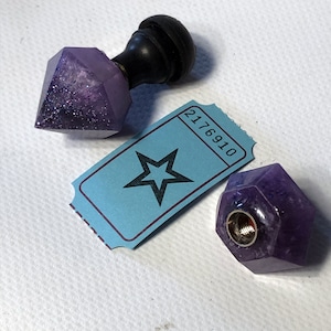 May include: Two purple amethyst crystal bottle stoppers. One is a diamond shape with a black wooden top. The other is a faceted shape with a silver metal top. The stoppers are on a white surface with a blue ticket that says "2176910".