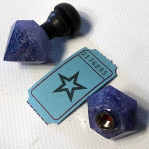 May include: Two purple and blue resin bottle stoppers with glitter. One stopper is shaped like a diamond and has a black wooden knob on top. The other stopper is shaped like a hexagon and has a gold metal screw top. A blue ticket with a star and the number 2176895 is in the background.