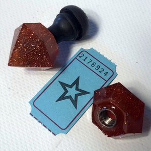 May include: Two red glitter bottle stoppers with a black top. One stopper is shaped like a diamond and the other is shaped like a hexagon. A blue ticket with the number 2176924 and a black star is in the background.