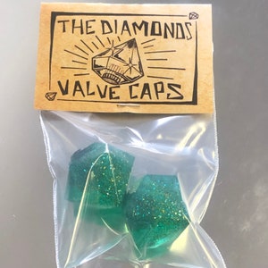 Puede incluir: Dos tapones de válvula en forma de diamante verde con purpurina en una bolsa de plástico transparente con una etiqueta de papel marrón que dice "The Diamonds Valve Caps".