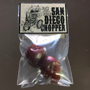 Peut inclure: Deux objets en forme de poing, rouge foncé, avec des accents argentés, emballés dans un sac en plastique transparent. Le sac porte une étiquette avec une illustration en noir et blanc d'une moto et les mots "Fuck the Rules" et "San Diego Chopper".