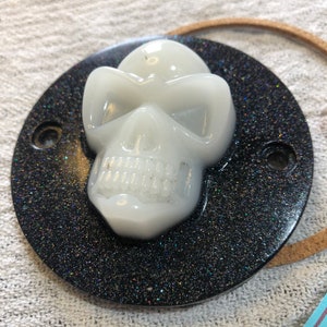 May include: A white skull with a black glitter background. The skull has a detailed face with eye sockets, a nose, and teeth. The background is a black circle with a sparkly finish.