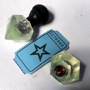 May include: Two clear green glass bottle stoppers with a star-shaped ticket in the background. The ticket has the number 2176948 printed on it.