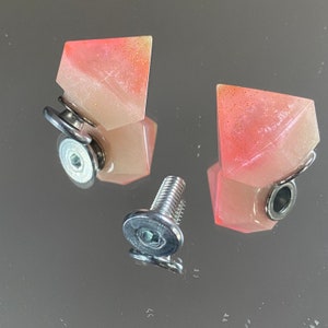May include: Two pink and white resin diamond-shaped drawer pulls with silver metal hardware. The drawer pulls are attached to a silver metal screw.