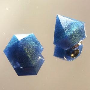 Può includere: Due anelli geometrici sfaccettati in resina blu con una lucentezza metallica. Un anello è in posizione verticale, con una fascia argentata e un centro cavo. L'altro anello è sdraiato su un lato. Lo sfondo è una superficie riflettente.