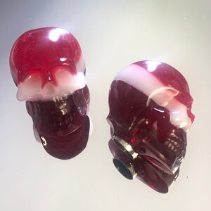 May include: Two red and white skull-shaped bottle stoppers. The stoppers are made of glass and have a shiny finish.