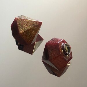 Può includere: Due ornamenti geometrici in resina in una finitura rosso scuro e glitter dorato. Un ornamento ha un'apertura rotonda nella parte superiore.