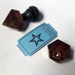 May include: Two amber glass bottle stoppers with a star-shaped ticket with the number 2176943. One stopper has a black rubber top and the other has a hexagonal shape.