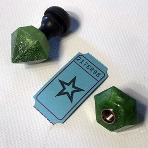 May include: Two green resin bottle stoppers with glitter, one shaped like a diamond and the other a hexagon. The stoppers are on a white surface with a blue ticket with a star and the number 2176898.