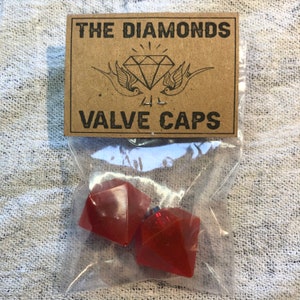 Può includere: Un sacchetto di plastica trasparente contenente due tappi per valvole a forma di diamante rosso. Il sacchetto ha un'etichetta di carta marrone su cui è scritto "THE DIAMONDS VALVE CAPS".