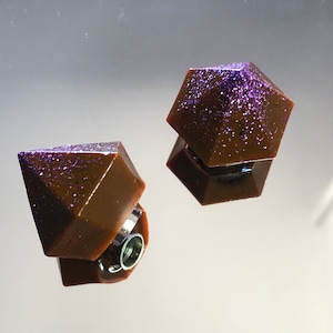 Puede incluir: Dos pendientes geométricos de color marrón oscuro con una parte superior de purpurina morada. Los pendientes se reflejan en una superficie espejada. Los pendientes tienen un poste plateado.