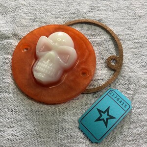 Può includere: Un pezzo rotondo in resina arancione con un design a teschio bianco, due piccoli fori e un anello in pelle marrone. È presente anche un biglietto blu con una stella e il numero 2176415. Gli oggetti sono su un tessuto grigio.